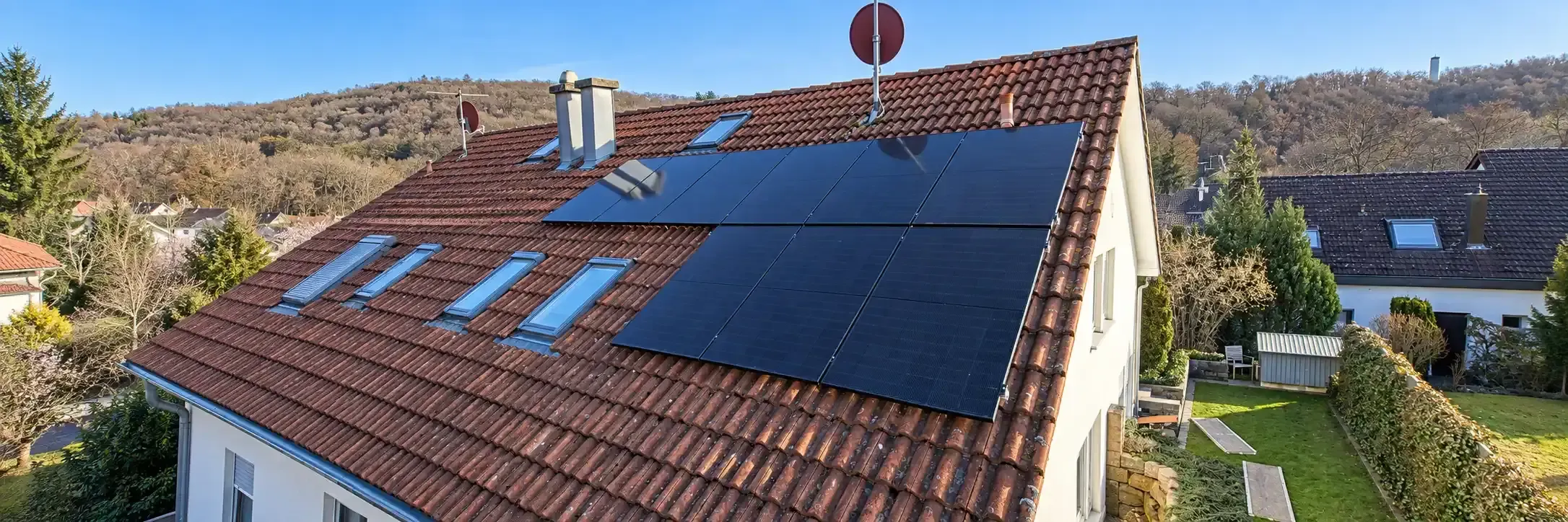 Eine Solaranlage auf dem Hausdach besteht aus mehreren großformatigen, dunkelblauen bis schwarzen PV-Modulen auf einem geneigten Ziegeldach. Die Anlage ist passend zwischen Dachfenstern, Schornstein und Dachrand angeordnet. Rund um das Einfamilienhaus sind Gärten, weitere Wohnhäuser und ein bewaldeter Hang zu sehen.