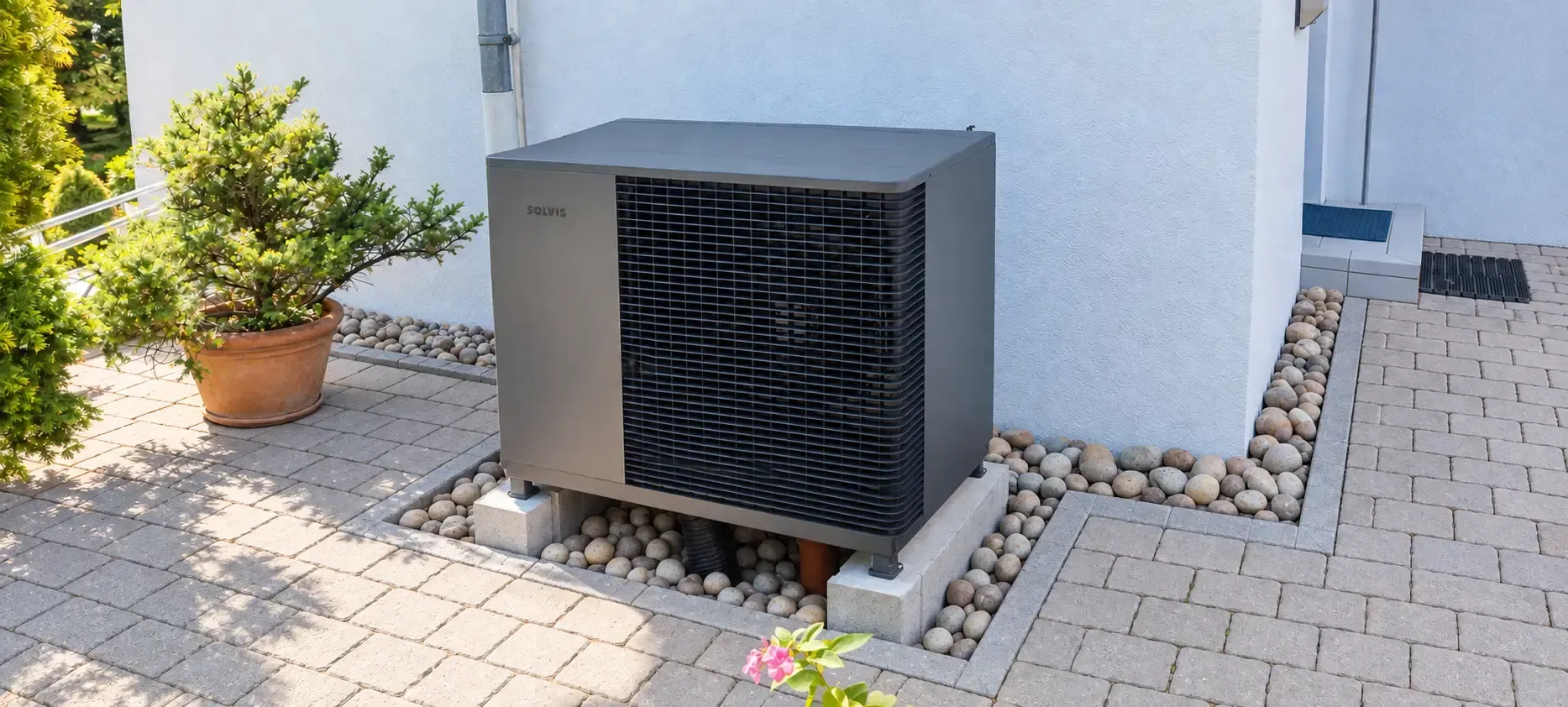 Eine Solvis Luft/Wasser-Wärmepumpe steht als Außeneinheit direkt an der Hauswand auf einem erhöhten Betonsockel. Das dunkelgraue Gerät ist von Kies eingefasst und fügt sich platzsparend in den gepflasterten Außenbereich des Wohnhauses ein.