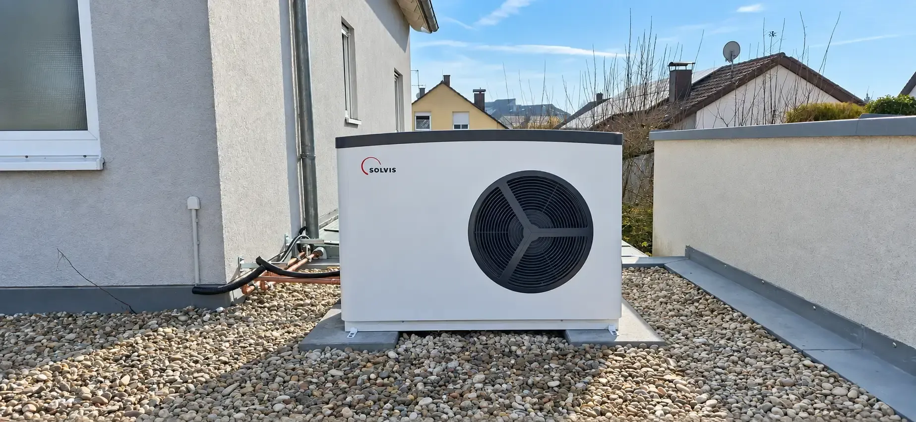 Eine **Solvis Luft/Wasser-Wärmepumpe** steht als Außeneinheit auf einer mit Kies bedeckten Fläche direkt neben dem Wohnhaus. Das weiße Gerät mit großem Ventilator ist auf einem festen Sockel montiert und über Leitungen mit dem Gebäude verbunden.