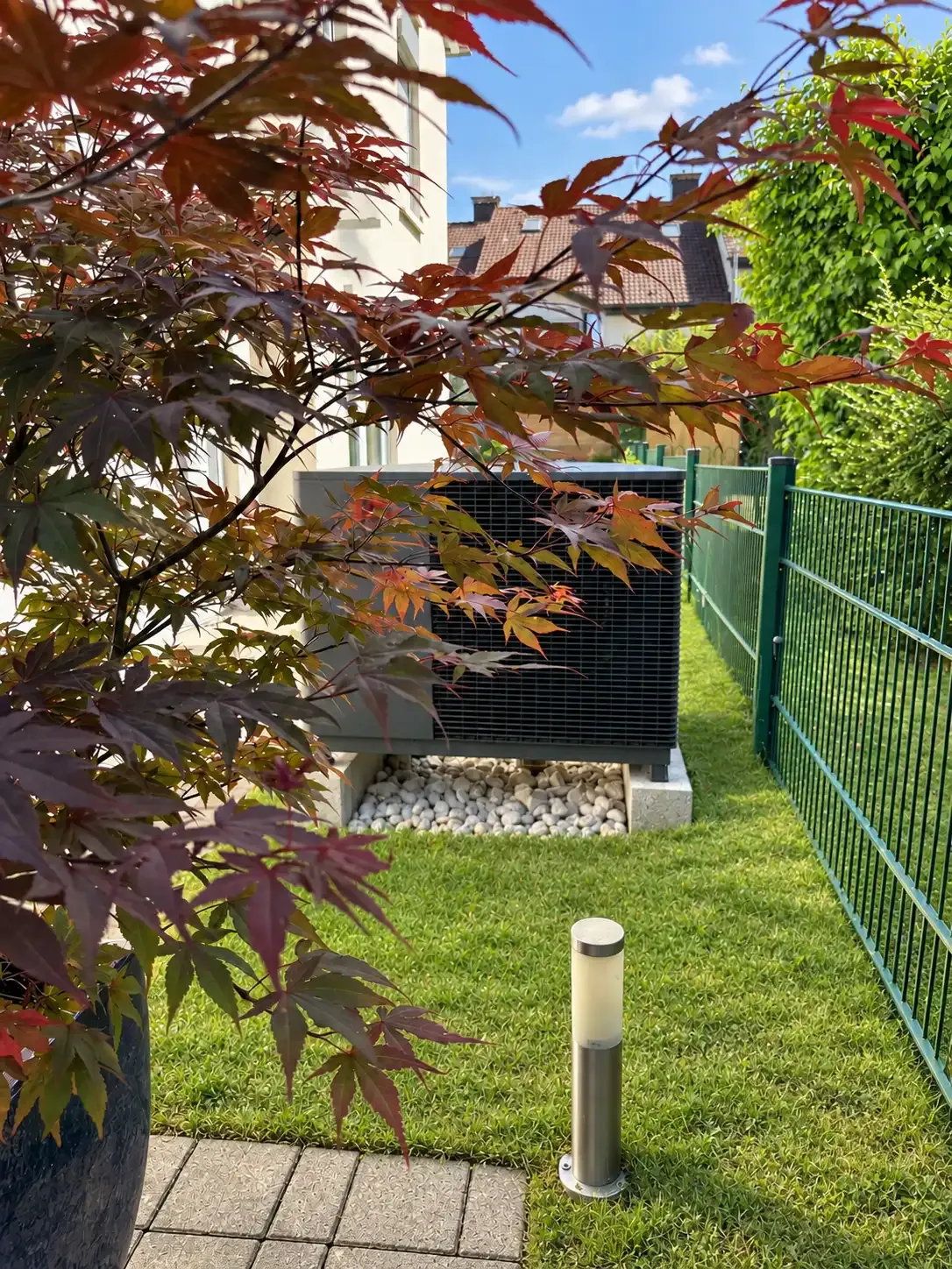Eine moderne Wärmepumpe im Garten steht auf einem Kiesfundament neben einem Wohnhaus und einem grünen Metallzaun. Das dunkelgraue Außengerät mit großem Lüftungsgitter wird im Vordergrund von den rotbraunen Blättern eines japanischen Ahorns eingerahmt. Die begrünte Umgebung zeigt, wie sich eine Wärmepumpe dezent in einen bestehenden Garten integrieren lässt.