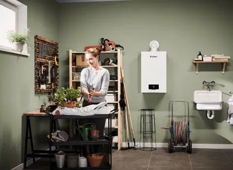 Eine Frau arbeitet an einem Pflanztisch in einer Werkstatt mit grüner Wand. Im Hintergrund hängt ein Vaillant-Gasbrennwertgerät neben Werkzeugen und Regalen.