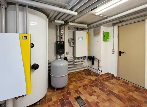 Eine Heizungsanlage mit großem Pufferspeicher ist in einem Technikraum installiert und über gedämmte Rohrleitungen mit weiteren Heizungs- und Regelungskomponenten verbunden. Neben dem Speicher sind ein Ausdehnungsgefäß, Steuerungseinheiten und verschiedene Armaturen der Wärmepumpenanlage zu sehen.