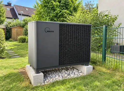 Die Wärmepumpe im Garten ist als freistehende Außeneinheit auf einem erhöhten Sockel neben einem Wohngebäude installiert. Auf der Vorderseite befindet sich ein großflächiges Schutzgitter vor dem Wärmetauscher, während darunter ein Kiesbett angelegt ist. Grüne Sträucher und Bäume integrieren die moderne Heiztechnik in die Gartenumgebung.