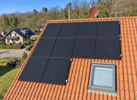 Eine Photovoltaikanlage auf einem Ziegeldach besteht aus mehreren dunkelblauen bis schwarzen PV-Modulen, die auf der geneigten Dachfläche angeordnet sind. Die Module sind rund um ein Dachfenster installiert; im Hintergrund sind weitere Wohnhäuser, Bäume und ein bewaldeter Hang zu sehen.