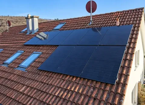 Eine Solaranlage auf dem Hausdach besteht aus mehreren großformatigen, dunkelblauen bis schwarzen PV-Modulen auf einem geneigten Ziegeldach. Die Anlage ist passend zwischen Dachfenstern, Schornstein und Dachrand angeordnet. Rund um das Einfamilienhaus sind Gärten, weitere Wohnhäuser und ein bewaldeter Hang zu sehen.