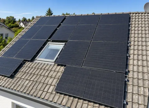 Eine großflächige Photovoltaikanlage mit zahlreichen schwarzen Solarmodulen bedeckt einen Großteil des geneigten Ziegeldachs eines Einfamilienhauses. Die Module sind rund um ein Dachfenster angeordnet und nutzen mehrere Bereiche der Dachfläche. Im Hintergrund sind weitere Wohnhäuser, Bäume und ein klarer blauer Himmel zu sehen.