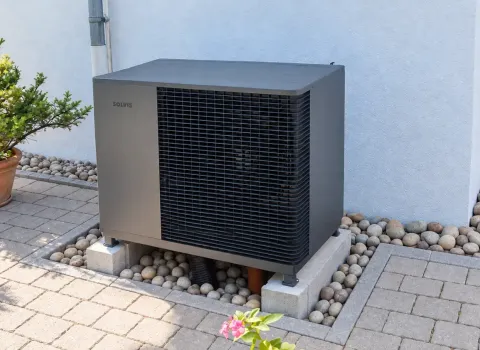 Eine Solvis Luft/Wasser-Wärmepumpe steht als Außeneinheit direkt an der Hauswand auf einem erhöhten Betonsockel. Das dunkelgraue Gerät ist von Kies eingefasst und fügt sich platzsparend in den gepflasterten Außenbereich des Wohnhauses ein.