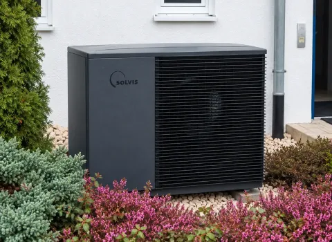 Eine **Solvis Luft/Wasser-Wärmepumpe** steht als Außeneinheit direkt an der weißen Hausfassade. Das dunkelgraue Gerät ist von hellen Kiessteinen, grünen Sträuchern und blühenden Pflanzen umgeben und harmonisch in den Vorgarten integriert.