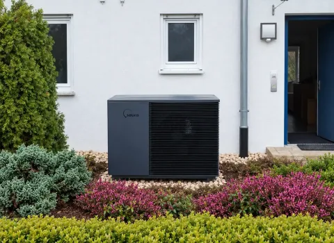 Eine Solvis Luft/Wasser-Wärmepumpe steht als Außeneinheit direkt an der weißen Fassade eines Einfamilienhauses. Die dunkelgraue Wärmepumpe ist sauber in den Vorgarten integriert und von grünen Sträuchern sowie blühenden Pflanzen umgeben.