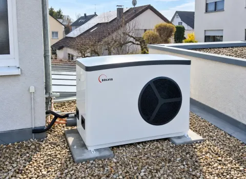 Eine Solvis Luft/Wasser-Wärmepumpe ist als Außeneinheit auf einer mit Kies bedeckten Dachfläche neben dem Wohnhaus installiert. Das weiße Gerät mit großem schwarzem Ventilator steht auf einem festen Sockel und ist über Leitungen mit dem Gebäude verbunden.