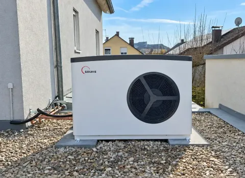 Eine **Solvis Luft/Wasser-Wärmepumpe** steht als Außeneinheit auf einer mit Kies bedeckten Fläche direkt neben dem Wohnhaus. Das weiße Gerät mit großem Ventilator ist auf einem festen Sockel montiert und über Leitungen mit dem Gebäude verbunden.