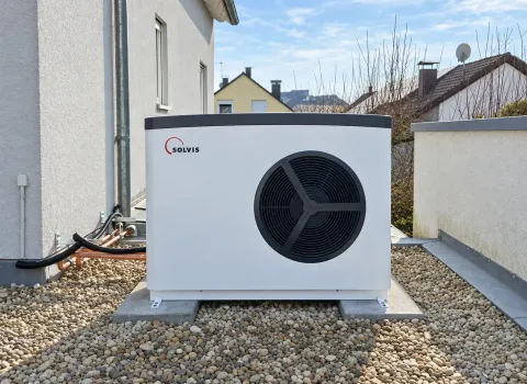 Eine Solvis Luft/Wasser-Wärmepumpe steht als Außeneinheit auf einer mit Kies bedeckten Fläche direkt neben dem Wohnhaus. Das weiße Gerät mit großem schwarzem Ventilator ist auf Betonplatten montiert und über Leitungen mit dem Gebäude verbunden.