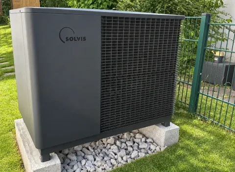 Eine dunkelgraue Solvis Wärmepumpe steht auf zwei Betonfundamenten mit einem Kiesbett in einem gepflegten Garten. Das Außengerät besitzt ein großflächiges schwarzes Lüftungsgitter und ist von Rasen, Sträuchern und Bäumen umgeben. Ein grüner Metallzaun verläuft unmittelbar neben der Anlage.