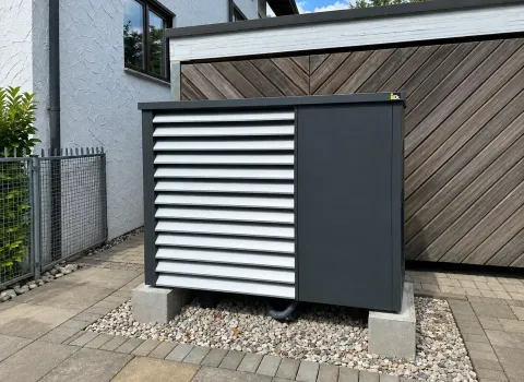 Eine **Wärmepumpe mit Lamellenverkleidung** steht auf Betonsockeln im gepflasterten Außenbereich neben einem Wohnhaus. Die dunkle Verkleidung mit hellen horizontalen Lamellen schützt die Außeneinheit und fügt sich optisch in die angrenzende Holzfassade und Hausumgebung ein.