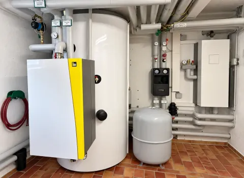 Eine Heizungsanlage mit Wärmepumpenspeicher steht in einem Technikraum und besteht aus einem großen zylindrischen Pufferspeicher, einem angeschlossenen IDM-System sowie einem Ausdehnungsgefäß. Zahlreiche gedämmte Rohrleitungen, Ventile und Regelungskomponenten verbinden die einzelnen Anlagenteile zu einem kompakten Heizsystem.
